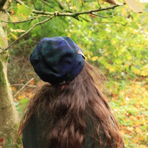 May include: A person wearing a dark green coat and a blue and green plaid hat. The hat has a small brown leather tag. The person has long, wavy brown hair. The background is a blurred view of a forest with green and yellow leaves.