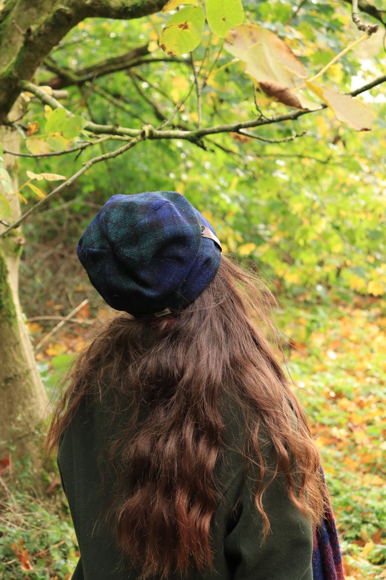May include: A person wearing a dark green coat and a blue and green plaid hat. The hat has a small brown leather tag. The person has long, wavy brown hair. The background is a blurred view of a forest with green and yellow leaves.