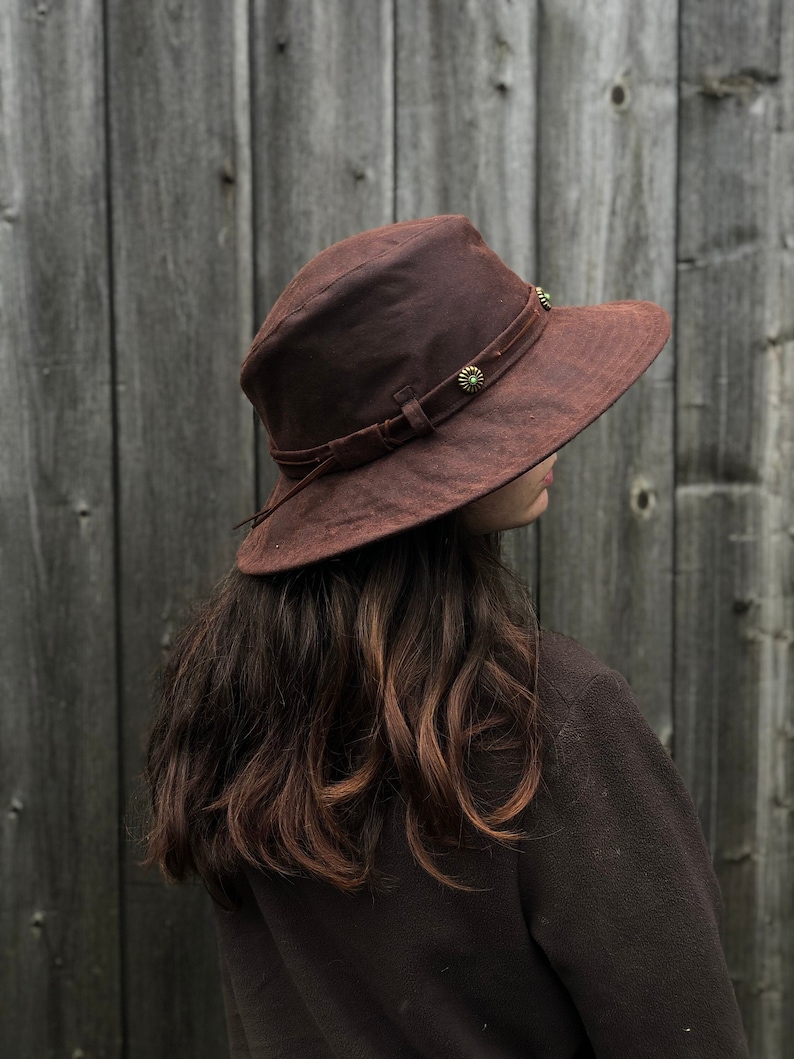 Puede incluir: Un sombrero marr&oacute;n de ala ancha con una banda de cuero y detalles met&aacute;licos decorativos. El sombrero est&aacute; hecho de un material suave y texturizado.