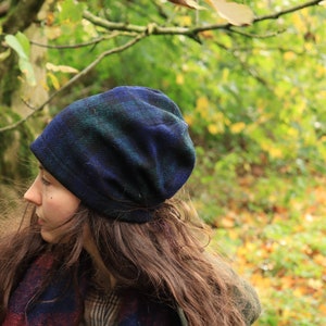 May include: A blue and green plaid beanie hat. The hat is made of a soft, textured fabric. The person is wearing a plaid scarf with red and blue accents and a plaid shirt. The background is a blurred view of a forest.