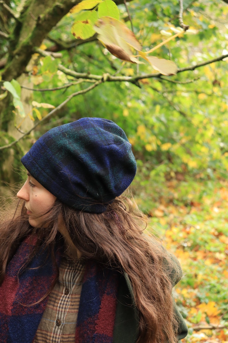 May include: A blue and green plaid beanie hat. The hat is made of a soft, textured fabric. The person is wearing a plaid scarf with red and blue accents and a plaid shirt. The background is a blurred view of a forest.