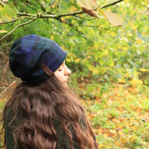 May include: A blue and green plaid hat with a brown leather strap detail. The hat is worn by a person with long brown hair, set against a backdrop of green and yellow foliage. The hat is a stylish accessory.