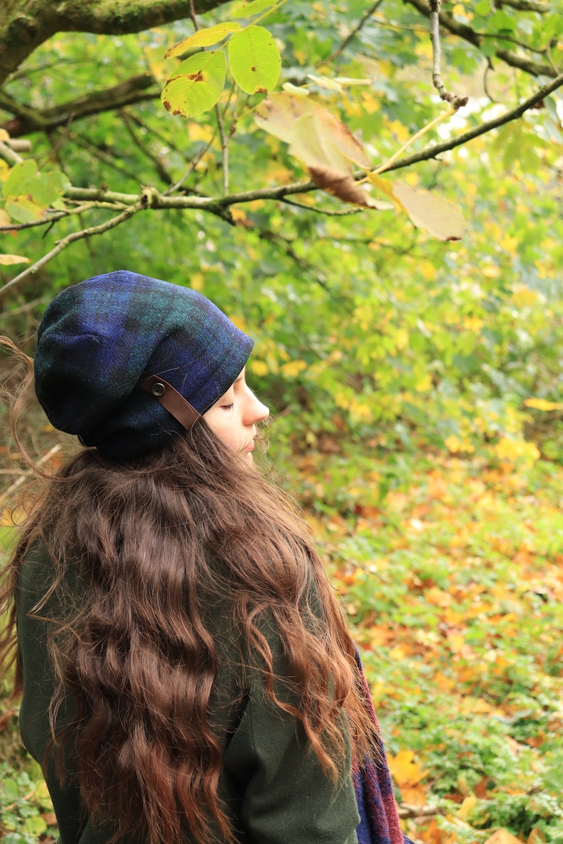May include: A blue and green plaid hat with a brown leather strap detail. The hat is worn by a person with long brown hair, set against a backdrop of green and yellow foliage. The hat is a stylish accessory.