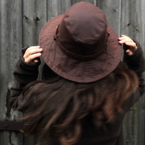 Puede incluir: Una persona con un sombrero marr&oacute;n de ala ancha y una chaqueta marr&oacute;n. El sombrero est&aacute; puesto bajo sobre la cabeza y el pelo es largo y oscuro.