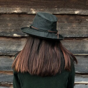 K&ouml;nnte beinhalten: Eine Person tr&auml;gt eine dunkelgr&uuml;ne Jacke und einen dunkelgr&uuml;nen Hut mit einem braunen Lederband. Die Person hat lange, dunkelbraune Haare und steht vor einer Holzwand.