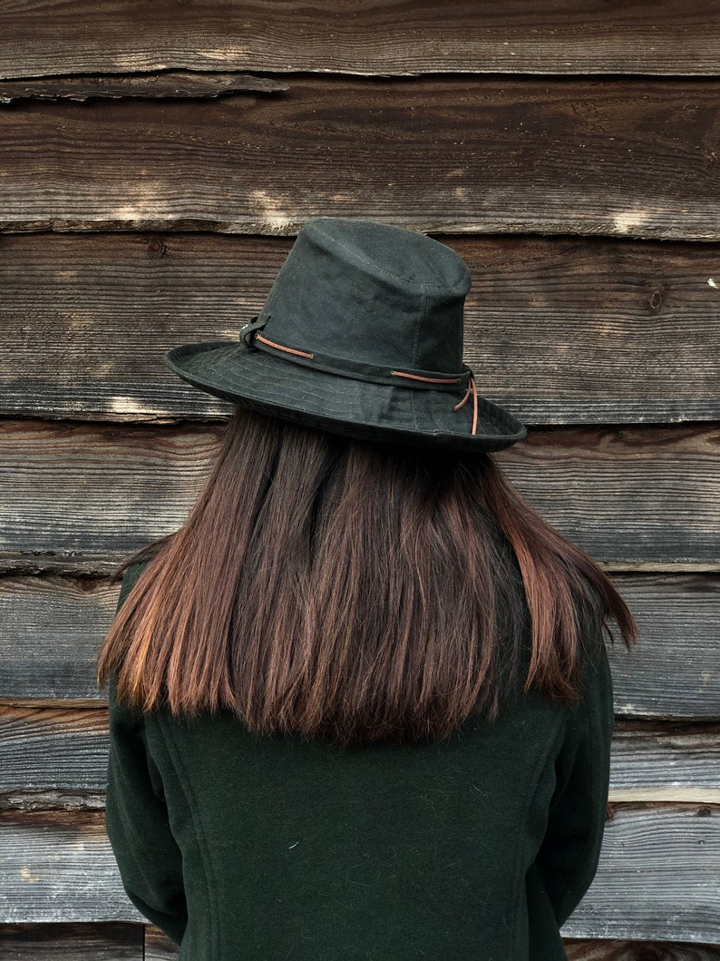 K&ouml;nnte beinhalten: Eine Person tr&auml;gt eine dunkelgr&uuml;ne Jacke und einen dunkelgr&uuml;nen Hut mit einem braunen Lederband. Die Person hat lange, dunkelbraune Haare und steht vor einer Holzwand.