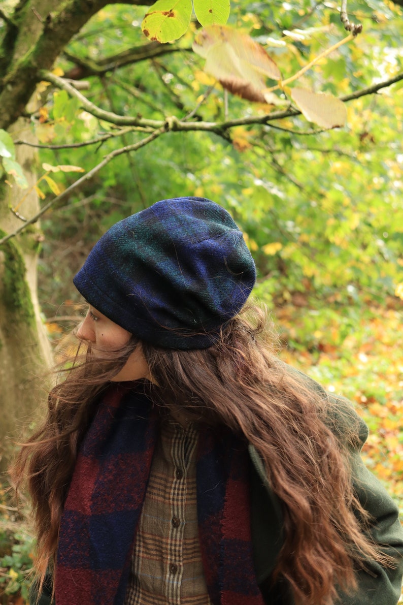 May include: A blue, green, and black plaid beanie hat and a matching scarf. The scarf is draped over a plaid shirt and a green coat. The background is a blurred view of a forest with green and yellow leaves.