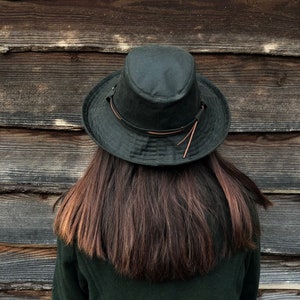 K&ouml;nnte beinhalten: Eine Person tr&auml;gt eine dunkelgr&uuml;ne Jacke und einen dunkelgr&uuml;nen Hut mit einem braunen Lederband. Die Person steht vor einer Holzwand.