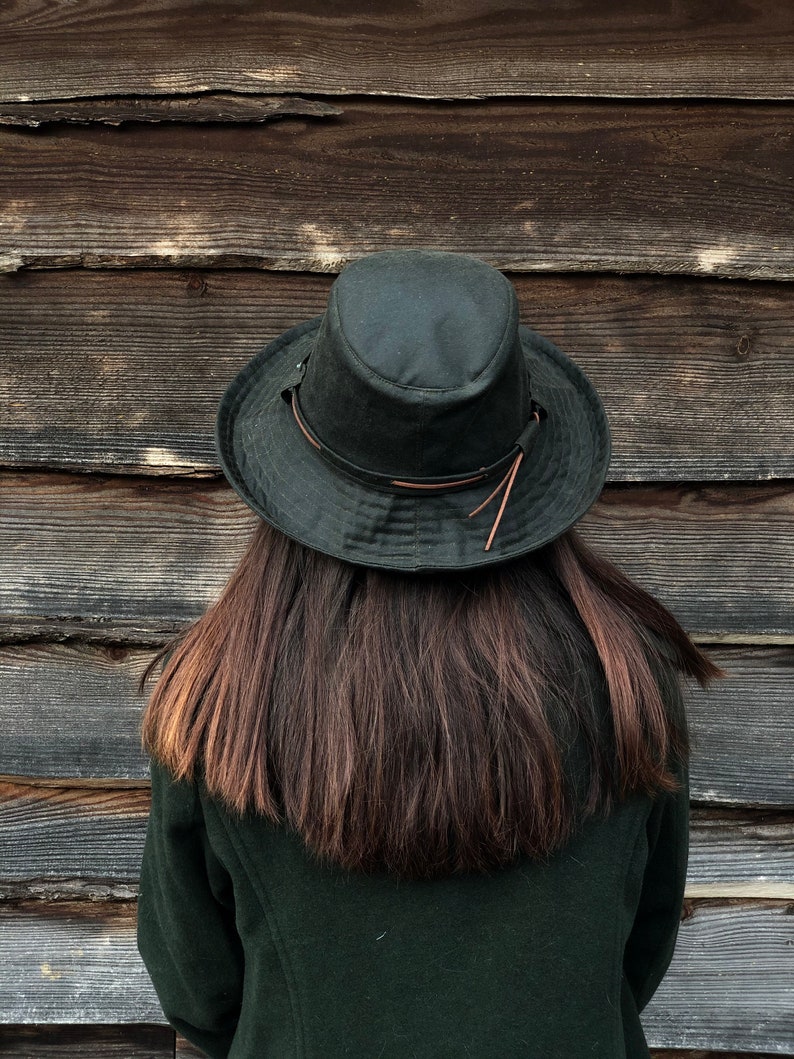 K&ouml;nnte beinhalten: Eine Person tr&auml;gt eine dunkelgr&uuml;ne Jacke und einen dunkelgr&uuml;nen Hut mit einem braunen Lederband. Die Person steht vor einer Holzwand.
