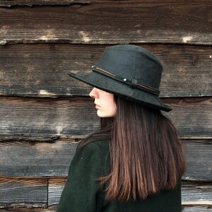 K&ouml;nnte beinhalten: Eine Person tr&auml;gt eine dunkelgr&uuml;ne Jacke und einen schwarzen Hut mit breiter Krempe und einem braunen Lederband. Die Person steht vor einer Holzwand.