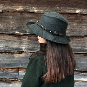 K&ouml;nnte beinhalten: Eine Person tr&auml;gt eine dunkelgr&uuml;ne Jacke und einen schwarzen, breitkrempigen Hut mit einem braunen Lederband. Die Person steht vor einer Holzwand.
