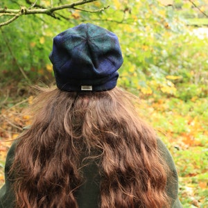 May include: A blue and green plaid beanie hat with a small white tag. The hat is worn by a person with long, wavy brown hair, and a dark green coat. The background is a blurred view of a forest with green and yellow leaves.