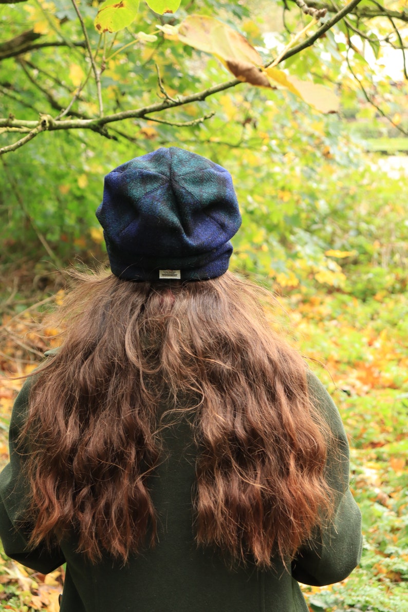 May include: A blue and green plaid beanie hat with a small white tag. The hat is worn by a person with long, wavy brown hair, and a dark green coat. The background is a blurred view of a forest with green and yellow leaves.
