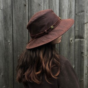 Puede incluir: Un sombrero marr&oacute;n de ala ancha con una banda de cuero y detalles met&aacute;licos decorativos. El sombrero est&aacute; hecho de una tela suave y texturizada.