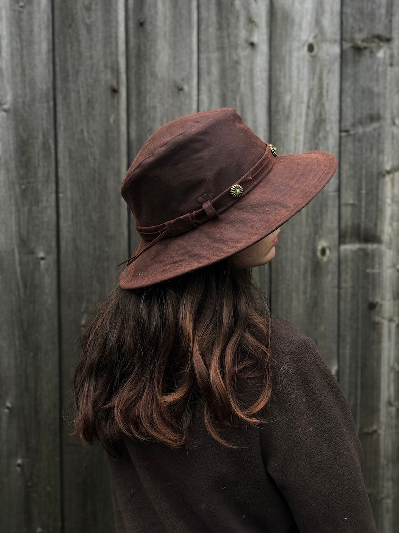 Puede incluir: Un sombrero marr&oacute;n de ala ancha con una banda de cuero y detalles met&aacute;licos decorativos. El sombrero est&aacute; hecho de una tela suave y texturizada.