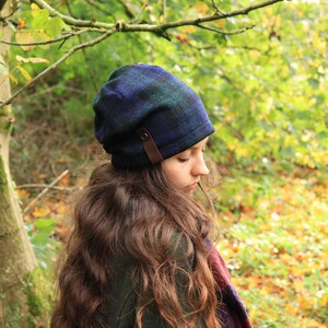 May include: A blue and green plaid beanie with a brown leather strap detail. The hat is worn by a person with long brown hair, and a plaid scarf is visible. The background features a forest with green and yellow foliage.