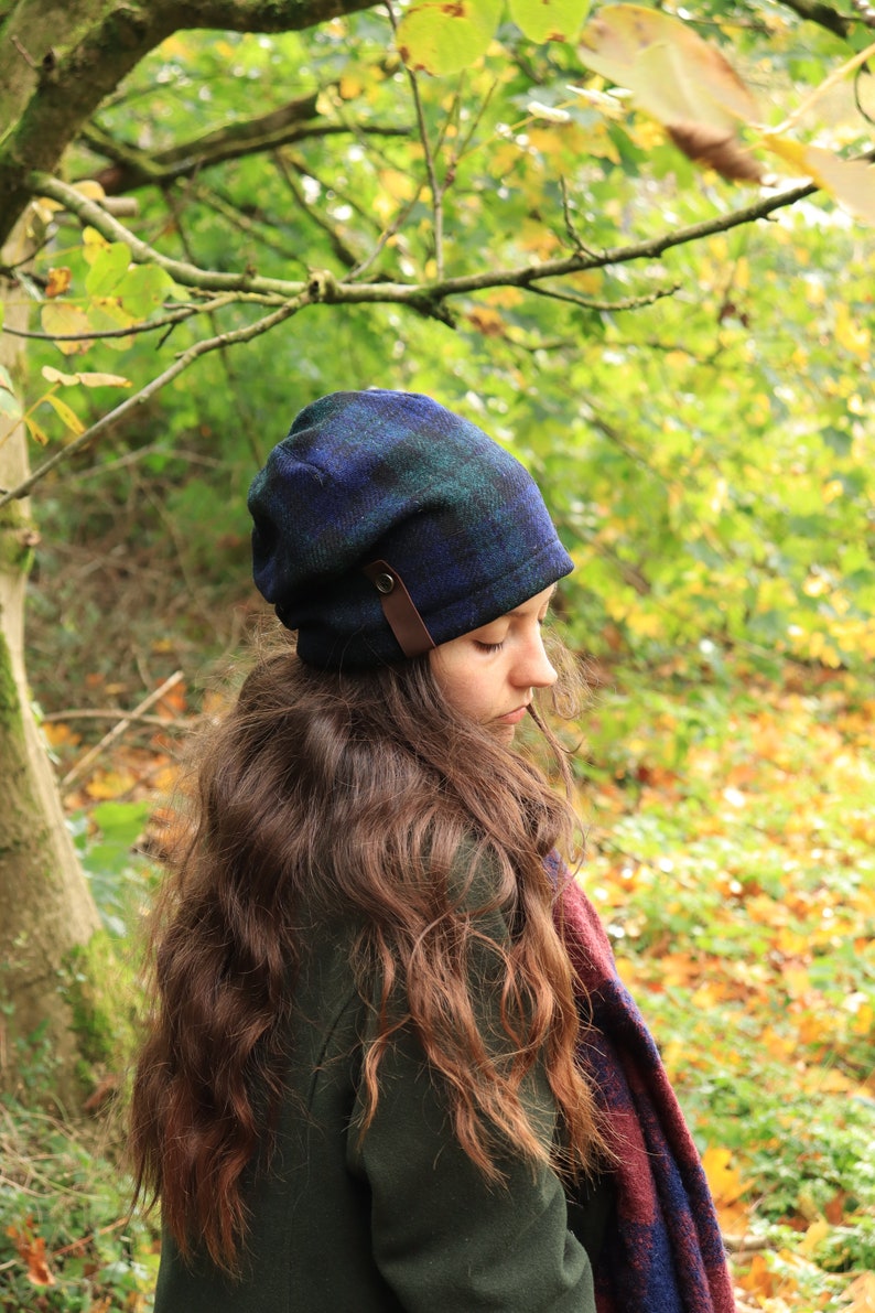 May include: A blue and green plaid beanie with a brown leather strap detail. The hat is worn by a person with long brown hair, and a plaid scarf is visible. The background features a forest with green and yellow foliage.