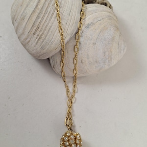 May include: A gold-toned necklace with a chain and a round pendant encrusted with small, clear crystals. The necklace is displayed on a white surface with two large, textured seashells.