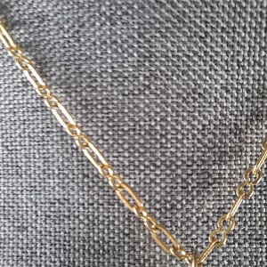 May include: Close-up of a gold-colored necklace with a delicate chain and a floral pendant. The pendant is adorned with small, clear gemstones. The necklace is displayed against a gray textured background.
