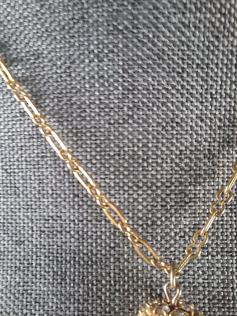 May include: Close-up of a gold-colored necklace with a delicate chain and a floral pendant. The pendant is adorned with small, clear gemstones. The necklace is displayed against a gray textured background.
