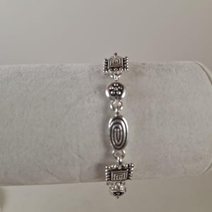 May include: Silver-toned bracelet with geometric and oval-shaped charms. The bracelet features square charms with intricate designs, a central oval charm with a spiral pattern, and a series of small, round charms. The bracelet is displayed against a neutral background.