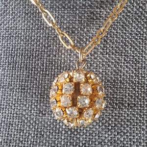 May include: A gold-toned necklace with a round pendant. The pendant is encrusted with clear, sparkling crystals, arranged in a grid pattern. The chain is a simple link style, and the background is a textured gray fabric.
