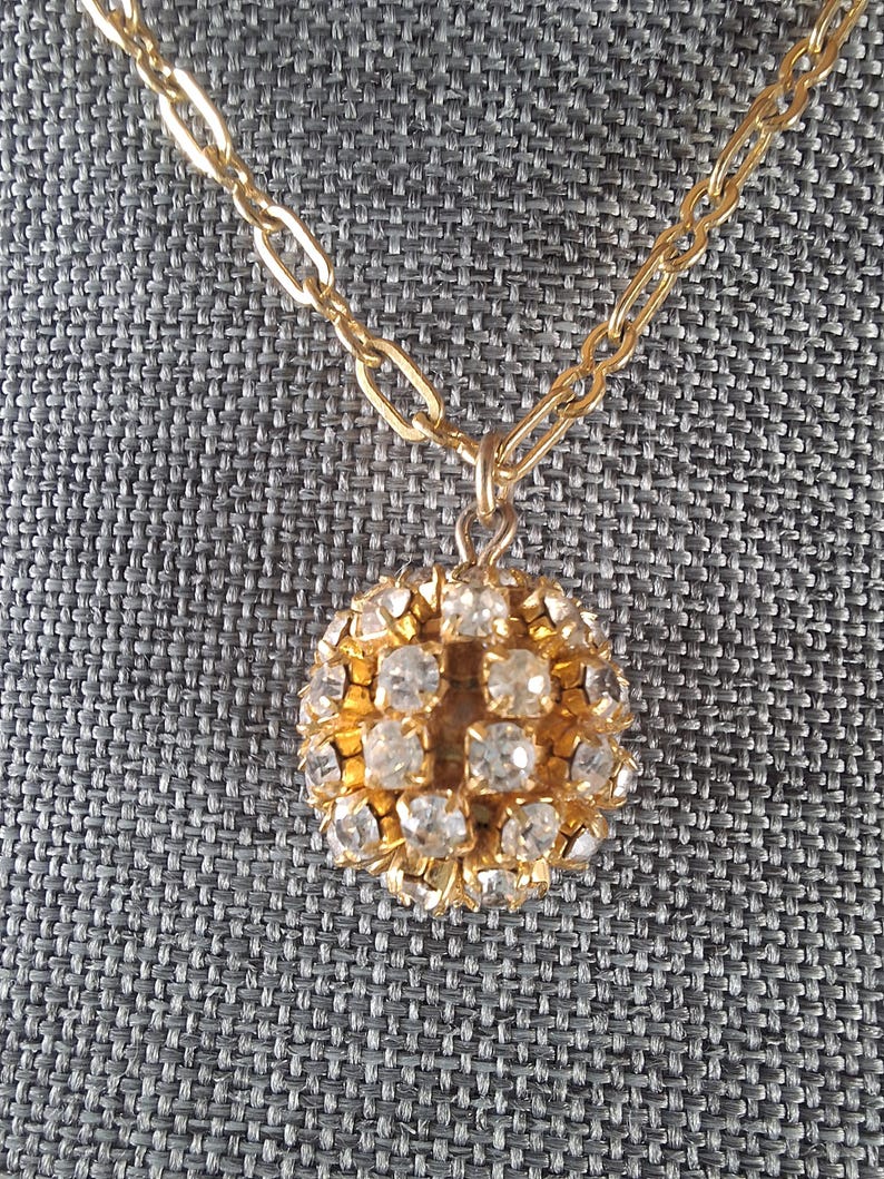 May include: A gold-toned necklace with a round pendant. The pendant is encrusted with clear, sparkling crystals, arranged in a grid pattern. The chain is a simple link style, and the background is a textured gray fabric.