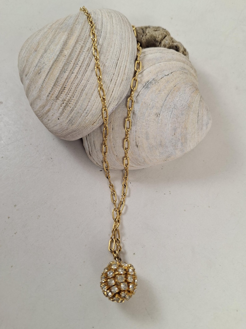 May include: A gold-toned necklace with a chain and a round pendant encrusted with small, clear crystals. The necklace is displayed on a white surface with two large, textured seashells.