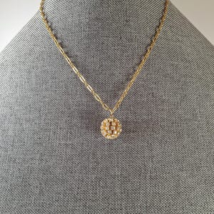May include: A gold-toned necklace with a delicate chain and a round pendant. The pendant is encrusted with small, sparkling stones, creating a dazzling effect. The necklace is displayed on a gray mannequin.