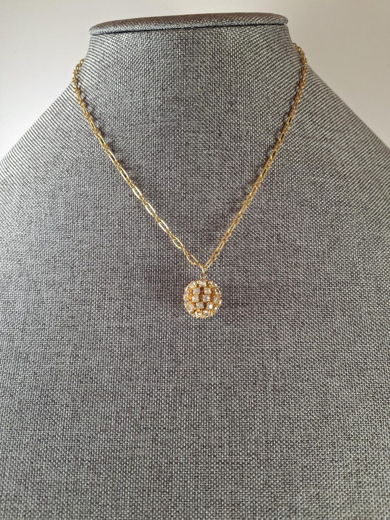 May include: A gold-toned necklace with a delicate chain and a round pendant. The pendant is encrusted with small, sparkling stones, creating a dazzling effect. The necklace is displayed on a gray mannequin.