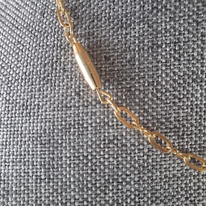 May include: Close-up of a gold-colored necklace with a chain link design. The necklace features a cylindrical gold-colored bar in the center. The background is a gray textured fabric.