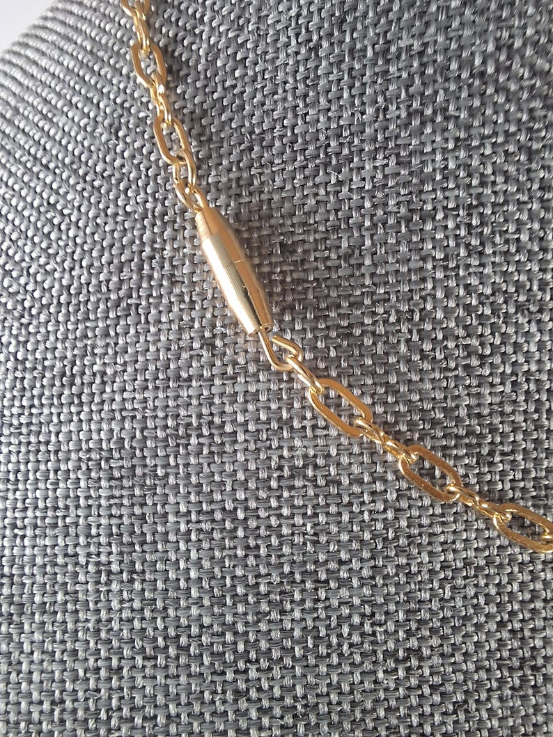May include: Close-up of a gold-colored necklace with a chain link design. The necklace features a cylindrical gold-colored bar in the center. The background is a gray textured fabric.