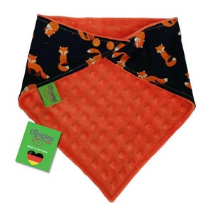 May include: A two-toned baby bib with a fox pattern on a navy blue background and a textured orange fabric. The bib has a snap closure and a green tag that says "Dimples Sew Happy" and "Made in Germany".