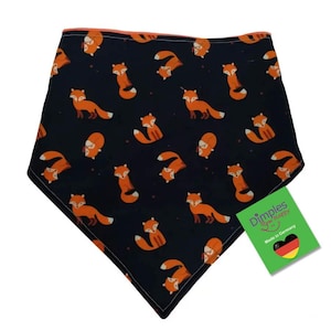 May include: A navy blue baby bib with a repeating pattern of orange foxes. The bib has a triangular shape and is trimmed with orange fabric. A green tag with the text "Dimples Sew Happy" and "Made in Germany" is attached.