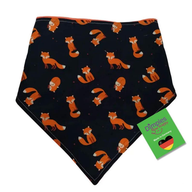 May include: A navy blue baby bib with a repeating pattern of orange foxes. The bib has a triangular shape and is trimmed with orange fabric. A green tag with the text "Dimples Sew Happy" and "Made in Germany" is attached.