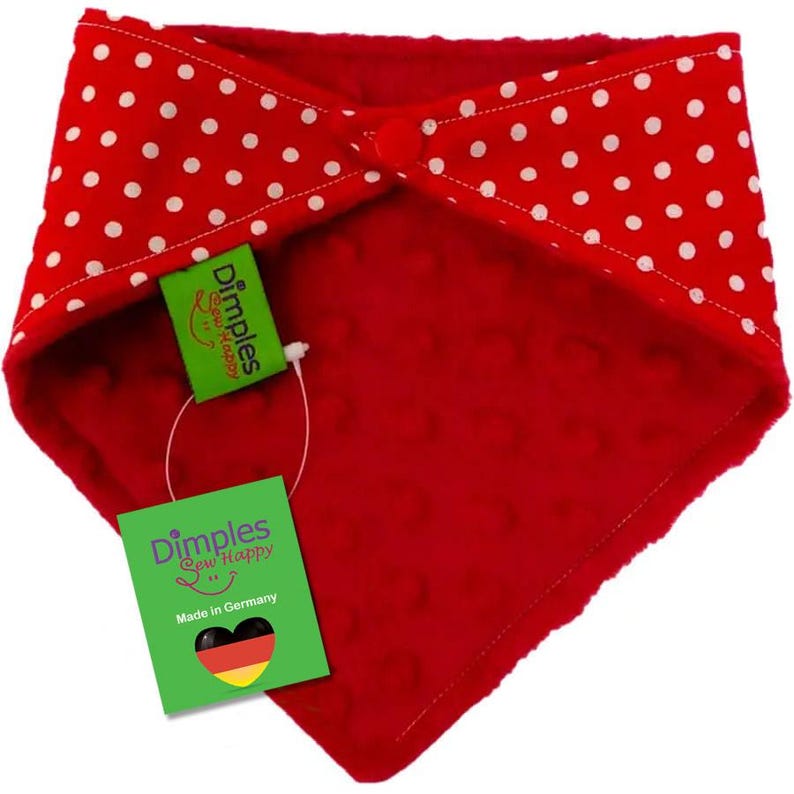 May include: A red baby bib with white polka dots on the top layer and a textured red fabric on the bottom layer. The bib has a button closure and a green tag that reads "Dimples Sew Happy" and "Made in Germany".