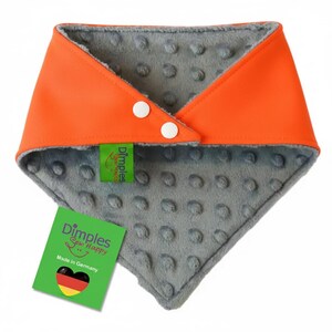 May include: Orange and gray baby bib with two white buttons. The bib has a soft, textured gray interior and a smooth orange exterior. A green tag reads "Dimples Sew Happy" and "Made in Germany."