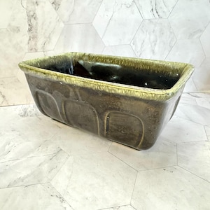 May include: A rectangular ceramic planter featuring a dark brown base color with a glossy finish. The upper rim is accented with a textured, light green drip glaze that contrasts with the darker body. The exterior sides display molded panels.