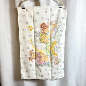 May include: A rectangular white fabric panel featuring a repeating pattern of small blue and pink flowers. The center displays a colorful illustration of three Cabbage Patch Kids dolls surrounded by flowers, a yellow butterfly, and a rainbow.