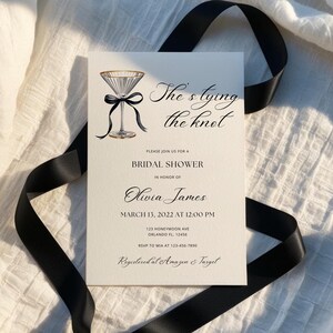 May include: A white bridal shower invitation with black calligraphy that reads "She's tying the knot". The invitation features a watercolor illustration of a martini glass with a black ribbon and bow. The invitation includes the bride's name, the date and time of the shower, the address, and RSVP information.
