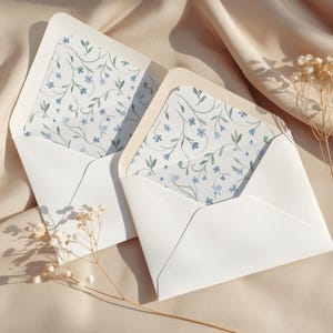 Op de afbeelding: Twee witte enveloppen met bloemenpatroonvoeringen, met blauwe bloemen en groene bladeren. De enveloppen zijn open en onthullen het delicate ontwerp. De scène speelt zich af op een beige stoffen achtergrond, met gedroogde bloemen.