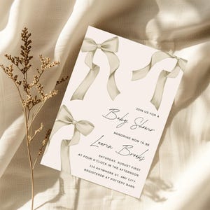 Puede incluir: Una invitación de baby shower rosa claro con un diseño de lazo beige. La invitación dice "Únete a nosotros para una Baby Shower en honor a la futura mamá Lauren Brooks. Sábado, 1 de agosto a las cuatro de la tarde. 123 Anywhere St. Any City. Registrado en Pottery Barn."
