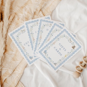 May include: A set of rectangular white cards with scalloped light blue borders, featuring floral wreaths and small illustrations of baby items like a rocking horse, teddy bear, and carriage. The text reads GIFTS & CARDS and CUST S D.
