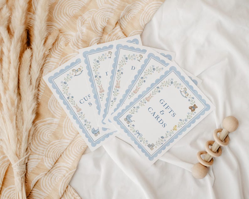 May include: A set of rectangular white cards with scalloped light blue borders, featuring floral wreaths and small illustrations of baby items like a rocking horse, teddy bear, and carriage. The text reads GIFTS & CARDS and CUST S D.