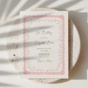May include: A pink and white invitation card for a 2nd birthday, featuring a scalloped border and floral accents. The text reads "2nd Birthday" and "Elizabeth Babu", with the date and time. The card is on a neutral surface.
