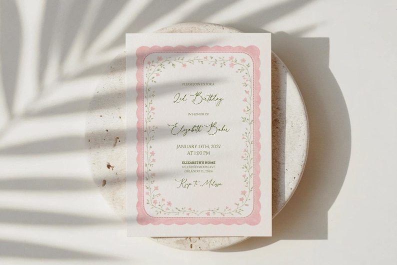 May include: A pink and white invitation card for a 2nd birthday, featuring a scalloped border and floral accents. The text reads "2nd Birthday" and "Elizabeth Babu", with the date and time. The card is on a neutral surface.