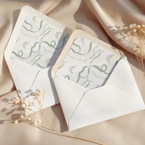 Könnte beinhalten: Zwei offene weiße Umschläge mit gemusterten Innenseiten mit grünen Bändern und floralen Akzenten. Die Umschläge liegen auf einer beigen Satin-Oberfläche, mit getrockneten Blumen im Vordergrund. Das Design ist zart und elegant.