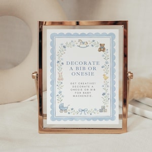May include: A rectangular rose gold frame containing a sign with a light blue scalloped border. The sign features floral illustrations, a rocking horse, a teddy bear, a rabbit, a duck, a stroller, and blocks with the text DECORATE A BIB OR ONESIE.