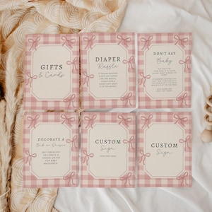 May include: A set of six baby shower signs with a pink gingham pattern and bow accents. The signs include text such as "Gifts & Cards", "Diaper Raffle", and "Don't Say Baby". A wooden rattle is also present.