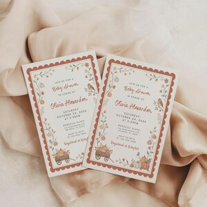 May include: Two rectangular baby shower invitation cards with scalloped rust-colored borders. The white cards feature illustrations of pumpkins, flowers, and a bird, with text reading Join us for a Baby Shower in honor of Olivia Alexander.